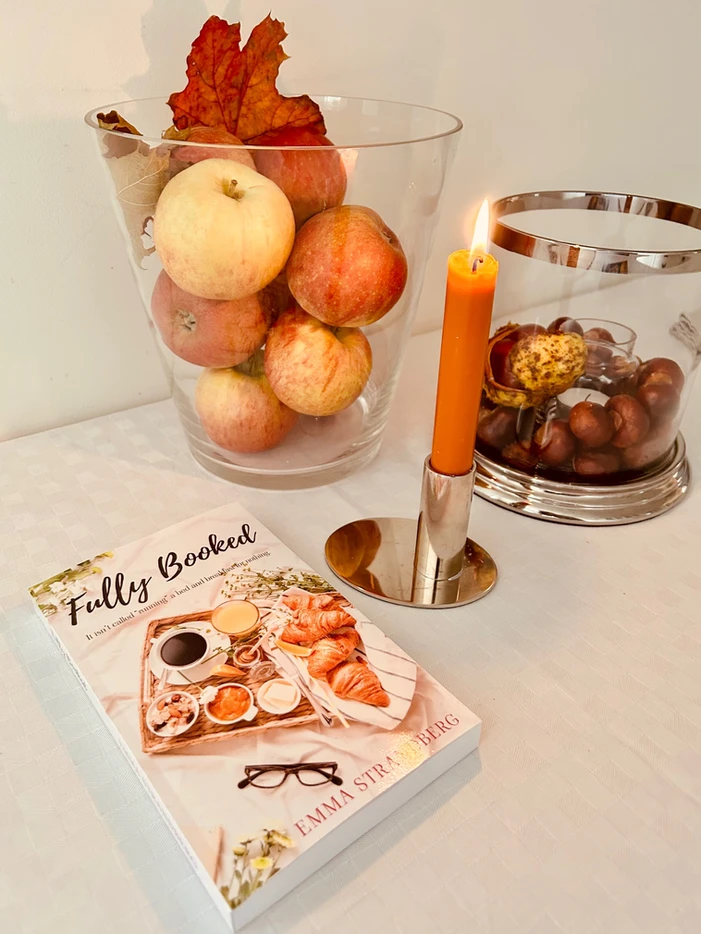 A cozy setting with apples in a glass vase, lit candle, nuts, and a book titled "Fully Booked" on a table.