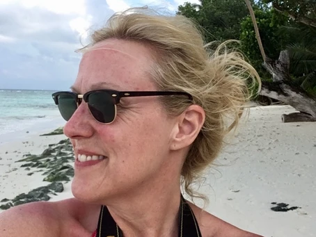 Blonde person on a beach with trees, sand, and ocean in the background.